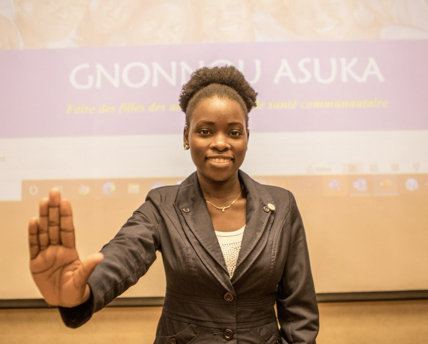 A young woman in a white shirt and black blazer extends her palm at the camera.