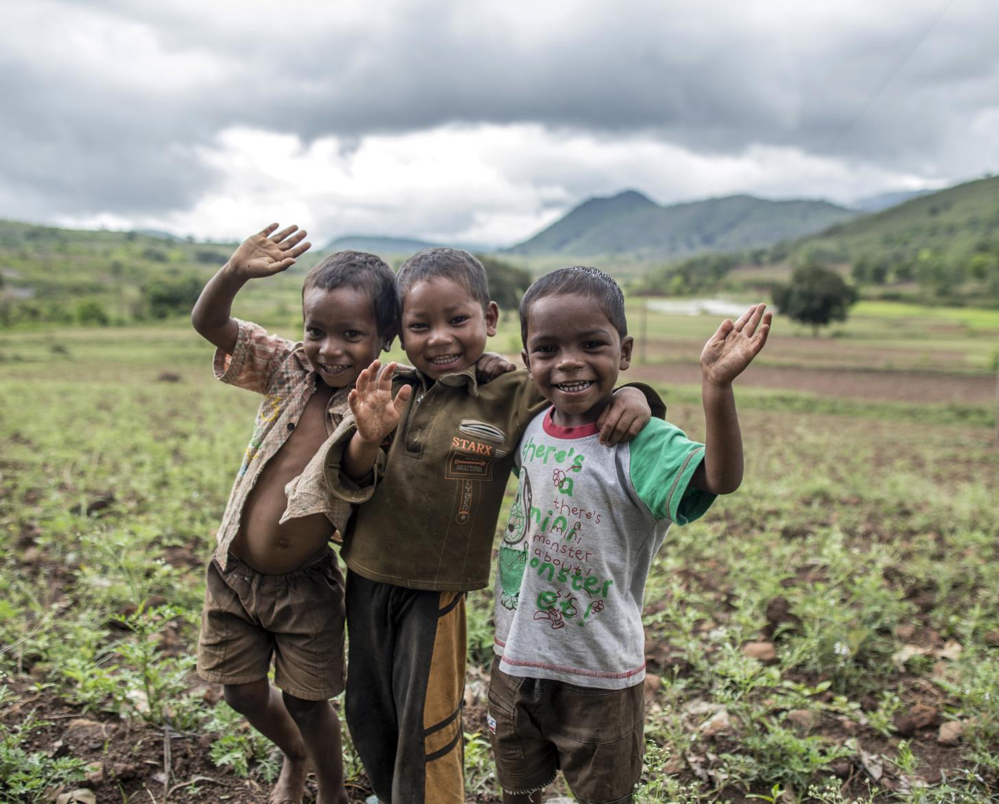 Three smiling boys in a field wave at the camera. 