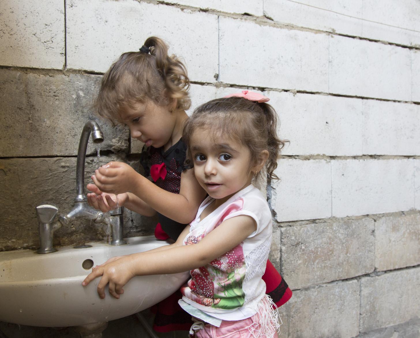 Two little girls washing their hands in an outdoor sink.