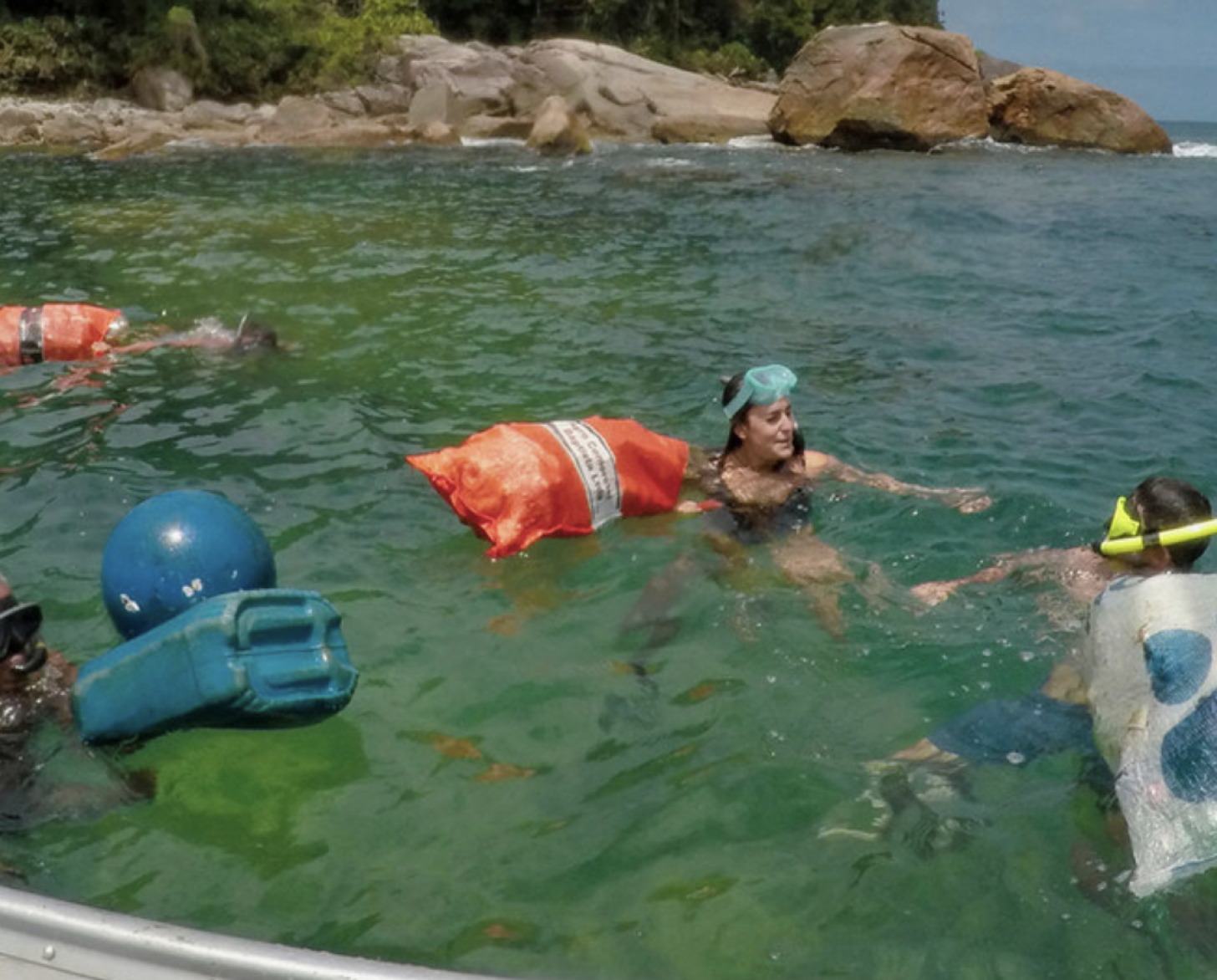 Several people swim in the ocean with large bags for waste collection and a floating plastic buoy.