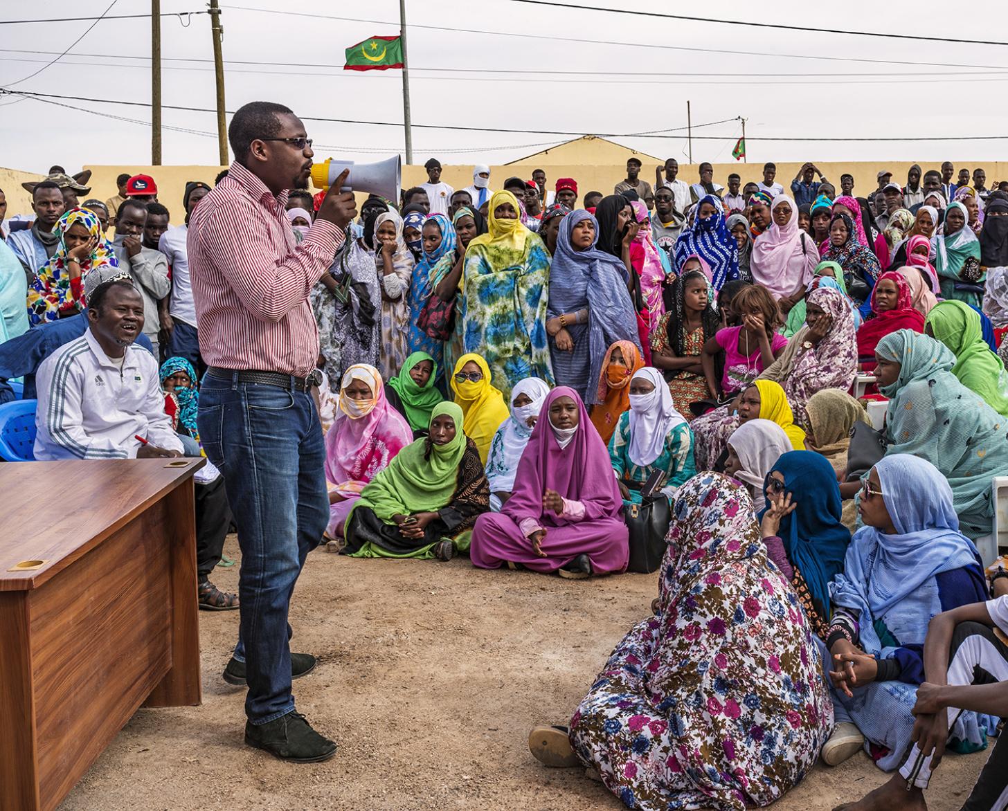 Muchas personas con atuendos multicolores se sientan alrededor de un hombre que habla en un escritorio.