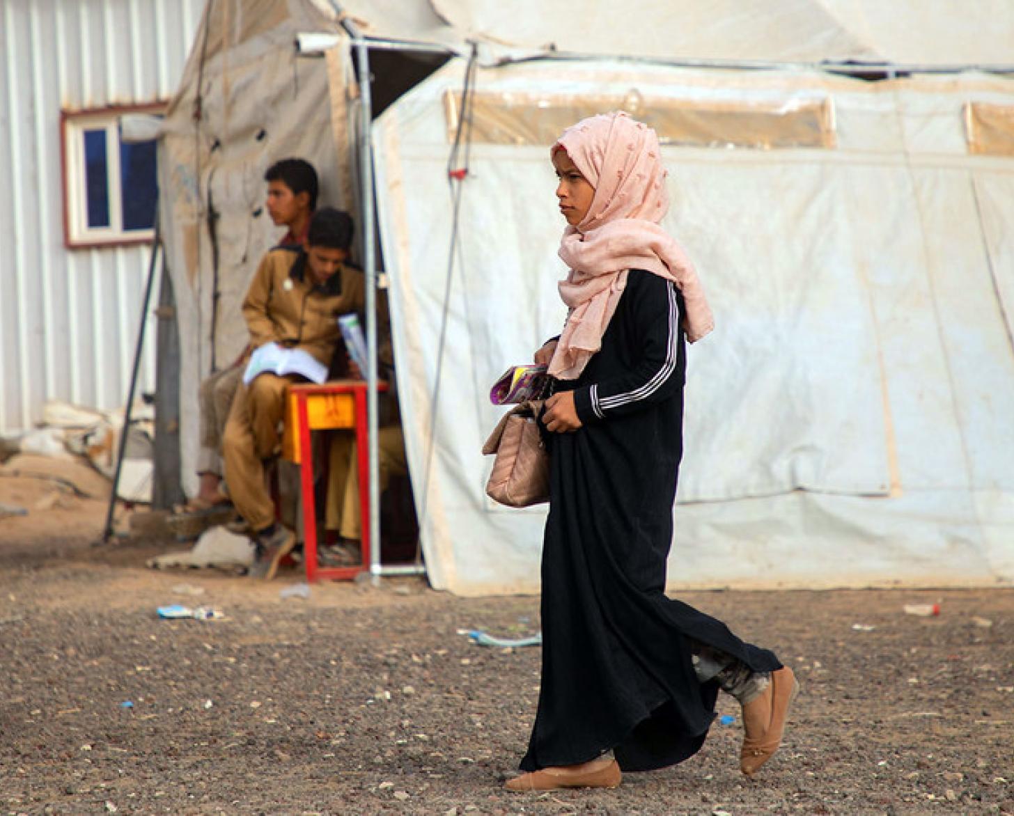 Une jeune fille yéménite vêtue de noir et portant un voile de couleur rose clair sur la tête se déplace dans un camp de réfugiés et passe devant une tente de couleur beige.