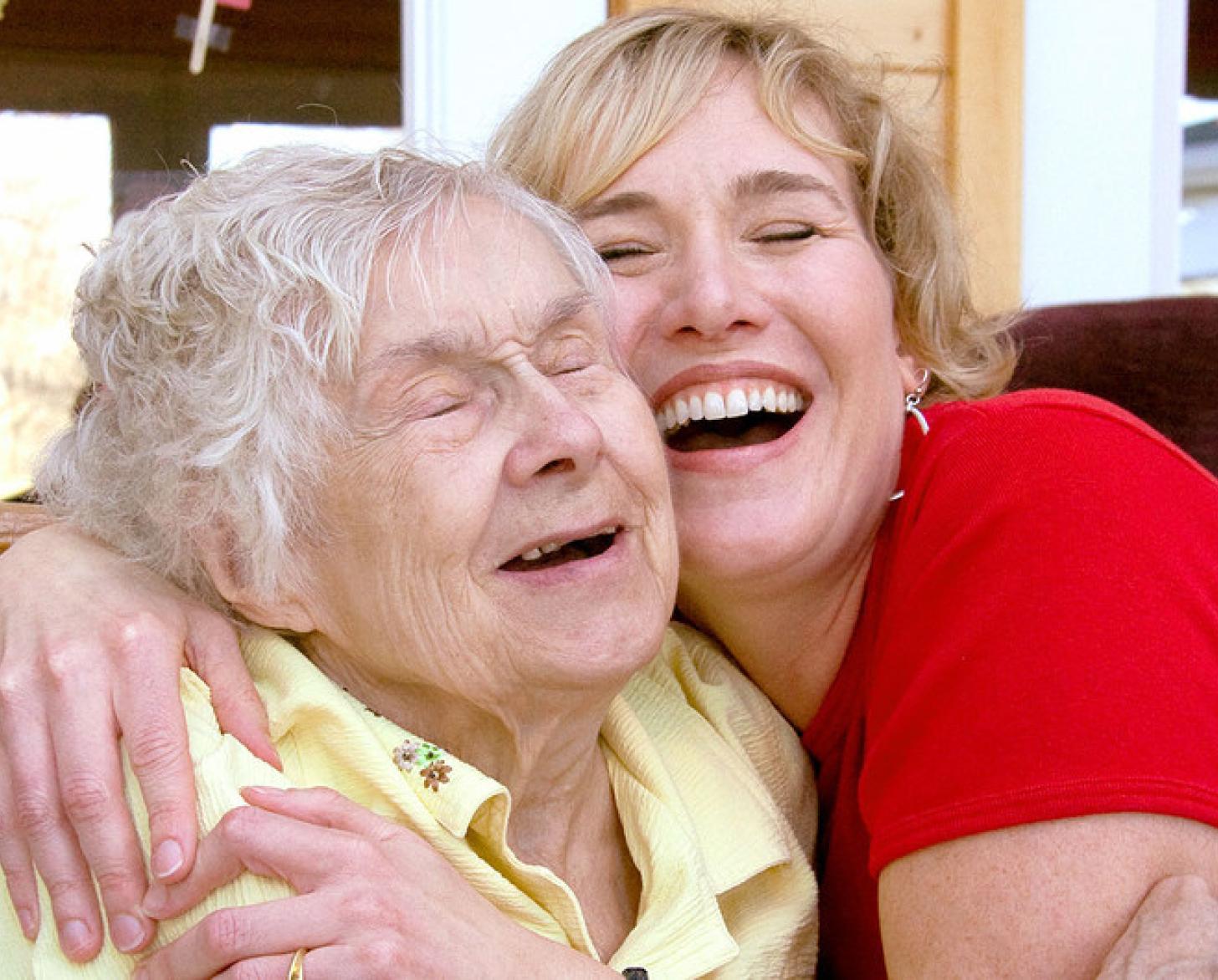 Une jeune femme blonde en t-shirt rouge et une femme âgée en polo jaune se prennent tendrement dans les bras l'une de l'autre, avec un grand sourire.