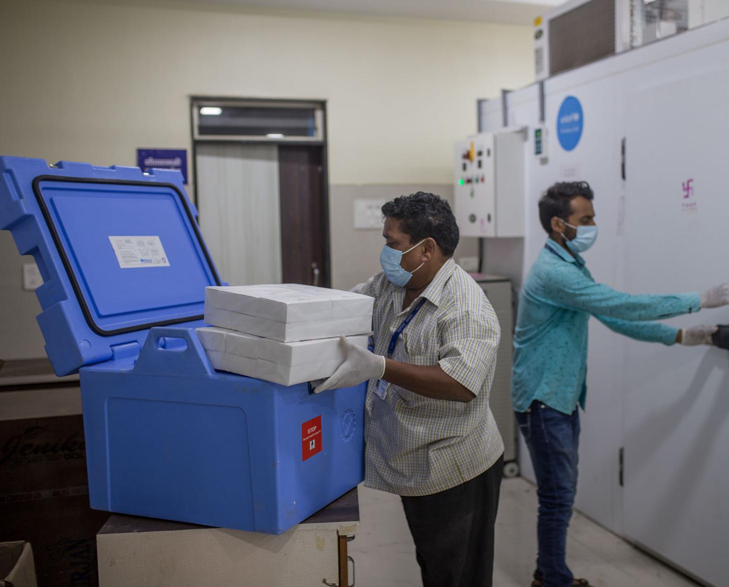 يقف اثنان من العاملين في مجال الرعاية الصحية بجوار ثلاجات كبيرة ويحمل أحدهما لقاحات كوفيد-19.