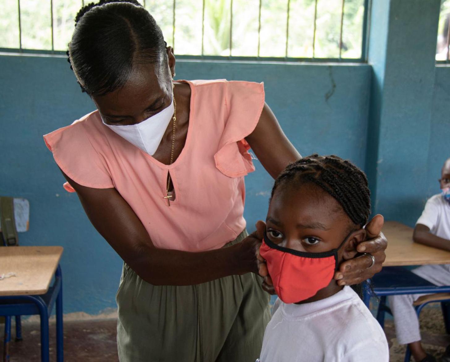 لقطة مقرّبة لمعلمة ترتدي كمامة في فصل دراسي تساعد طالبة في المرحلة الابتدائية على وضع كمامتها.