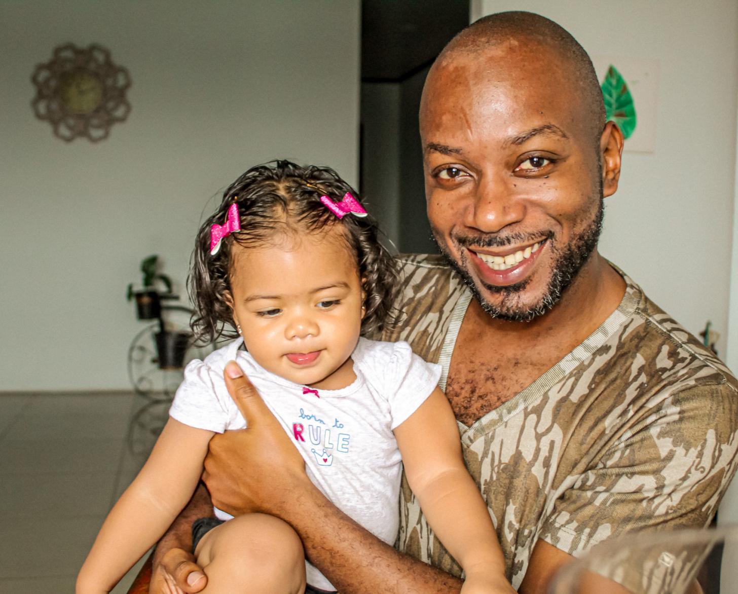 Un hombre está sentado en una habitación y sostiene en su regazo a un niño con barretina rosa.
