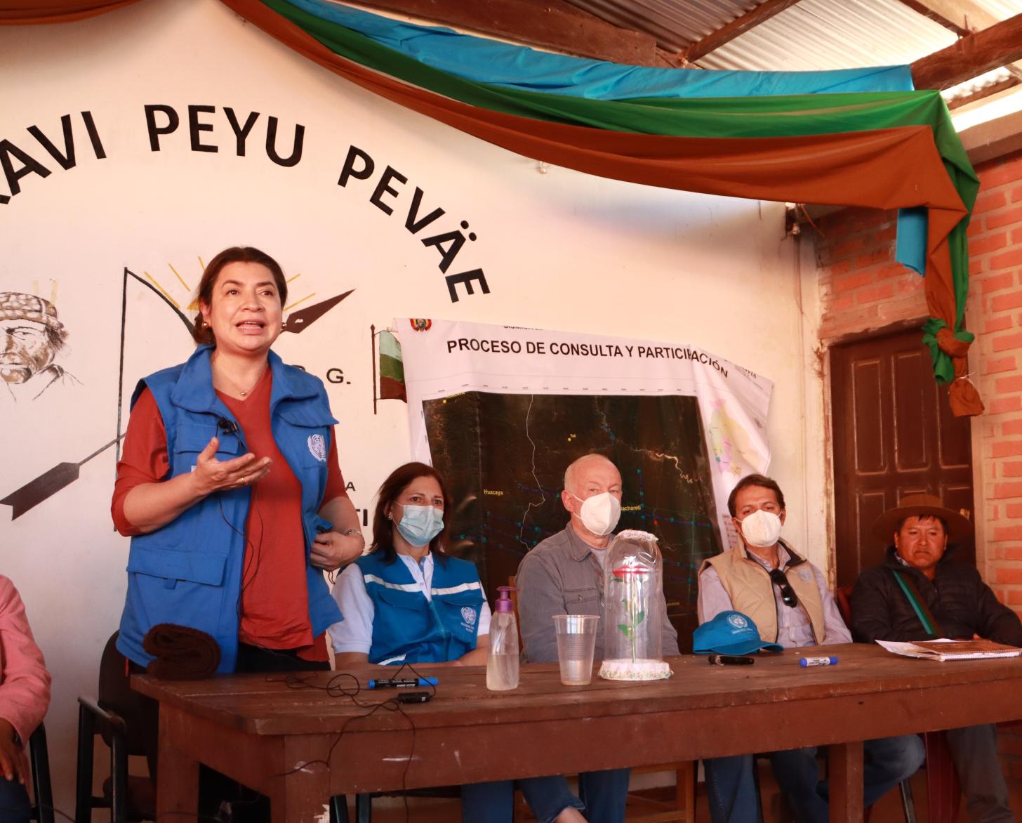 Un grupo de personas con mascarillas sentados frente a una audiencia, mientras Claudia Mojica, Coordinadora Residente de ONU Argentina se dirige a los presentes, durante la visita a la planta de transformación de productos de la agrobiodiversidad en Macharetí Bolivia.