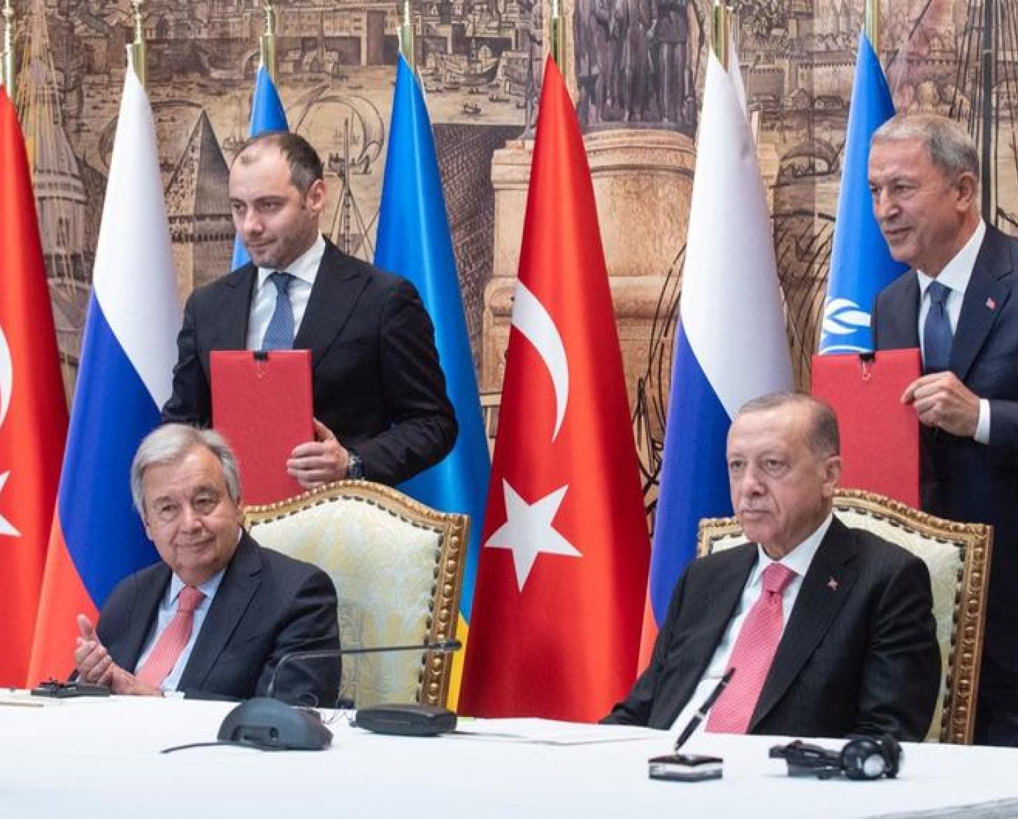 Two men in suits sit at a table with microphones ahead of them, with aides standing behind them. The table is adorned with Russian, Turkish, and UN flags.