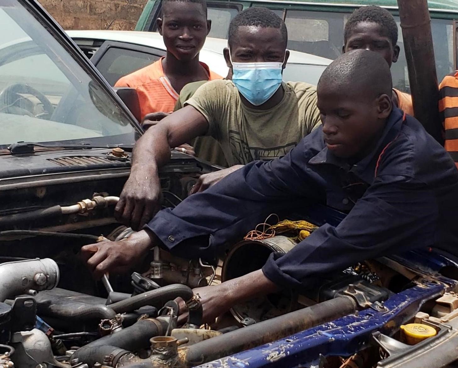 Un adolescent béninois vêtu d'une blouse de mécanien bleue et entouré de plusieurs de ses camarades est penché sur le moteur d'un véhicule et le répare.