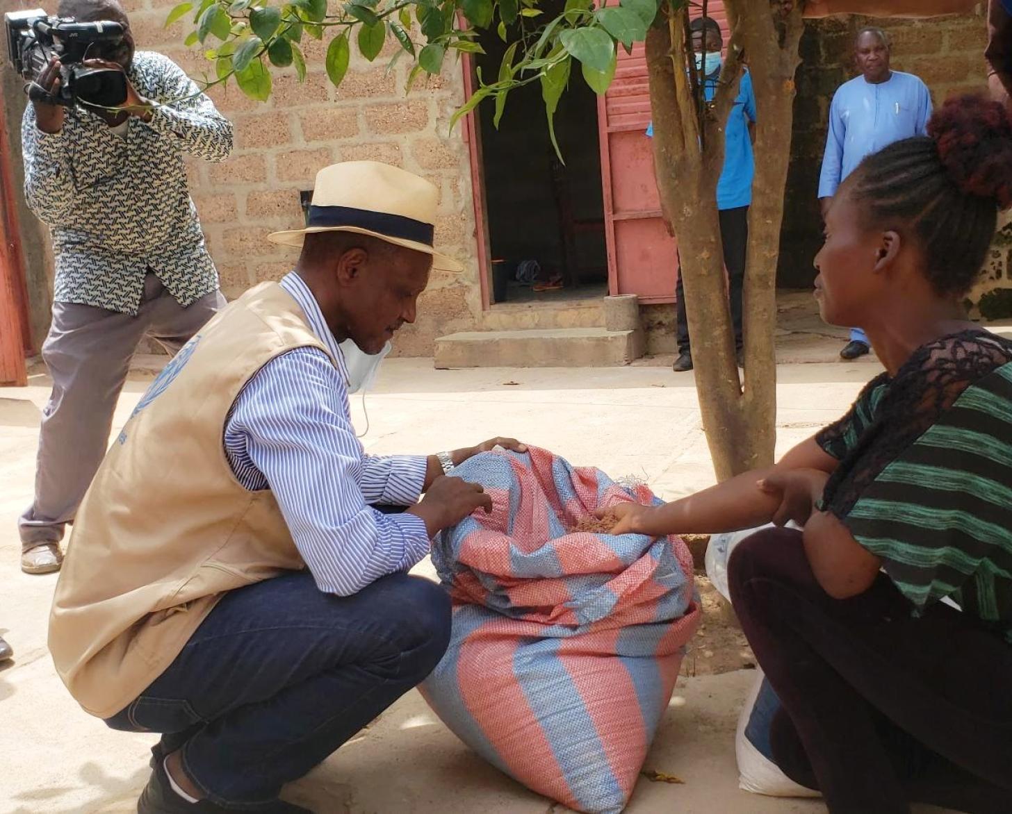 Une jeune femme béninoise et un homme portant un chapeau en paille sont accroupis de part et d'autre d'un grand un sac de provende, au pied d'un arbre, entourés de plusieurs autres personnes.