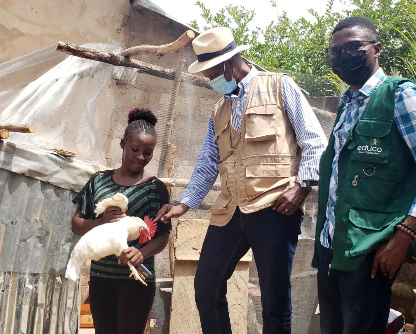 Une jeune femme et deux hommes se tiennent debout devant une habitation recouverte de toiles et de taules. L'homme qui se tient au milieu tend la main vers le coq que tient la jeune femme entre les mains. L'autre homme regarde en direction de la caméra.