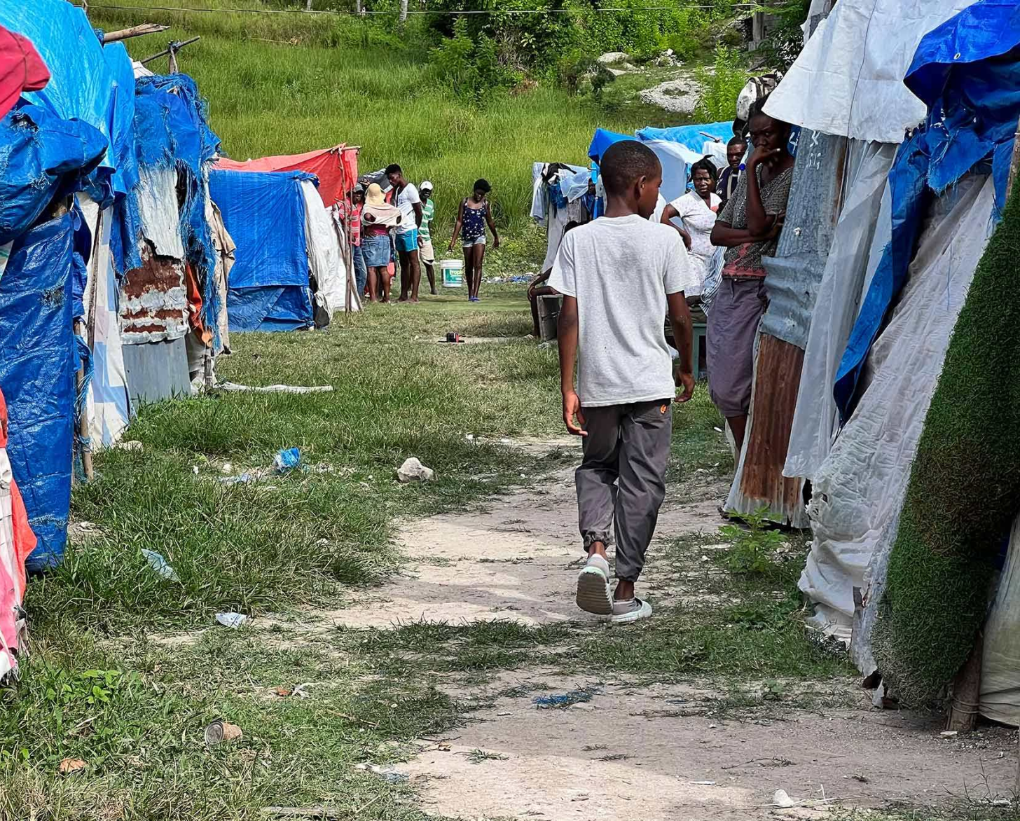 Un joven es fotografiado de espaldas caminando por un callejón bordeado por dos hileras de tiendas de campaña hechas con lonas de diferentes colores.