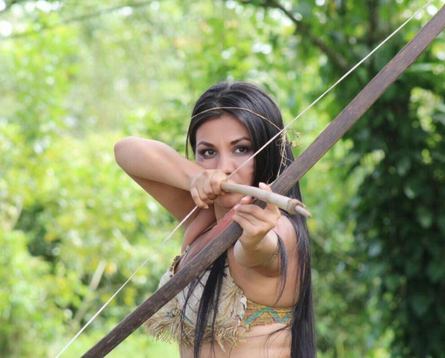 En medio de un paraje natural, una mujer enfocada en apuntar con un arco y una flecha.