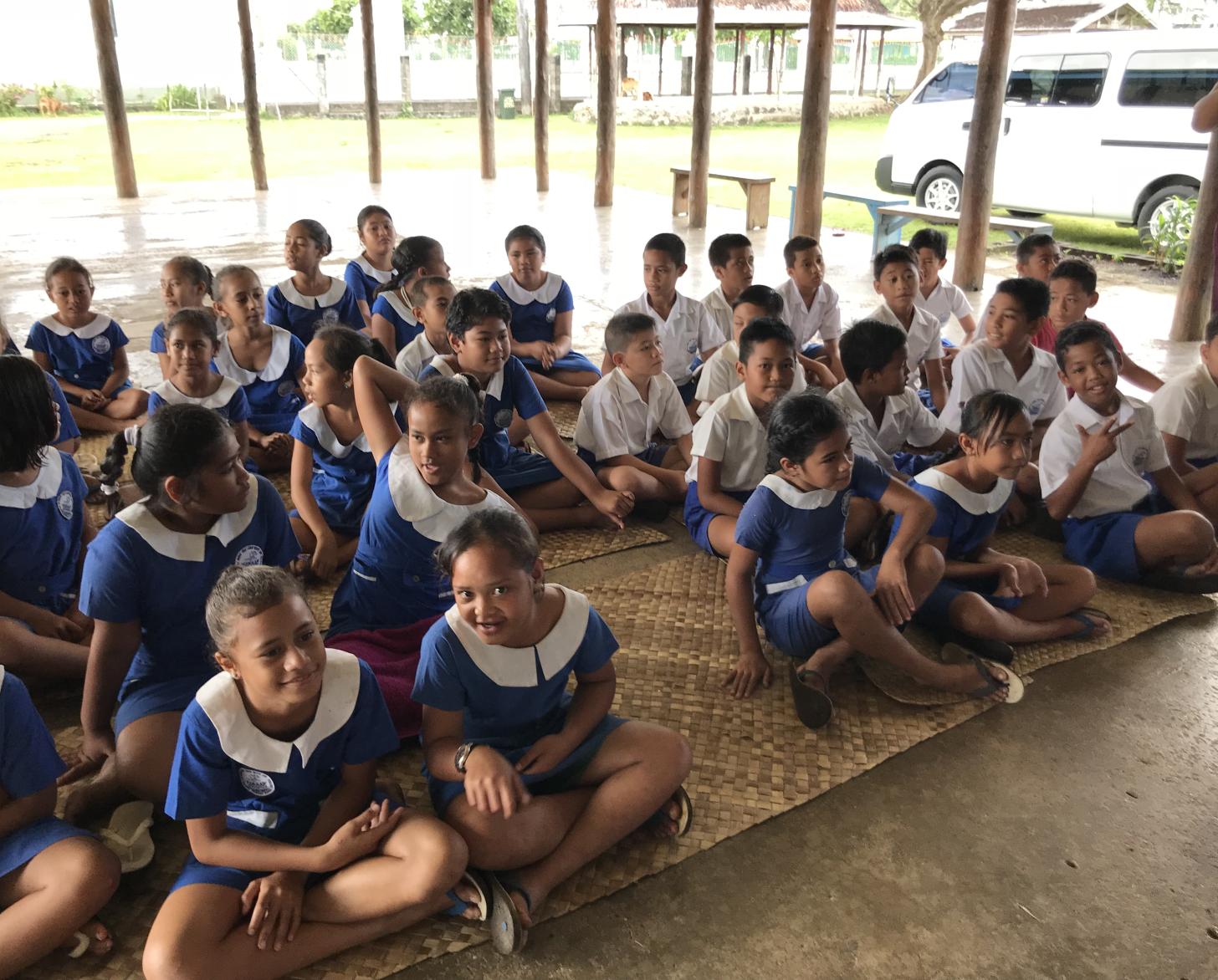 أطفال يرتدون الزي المدرسي يجلسون على حصائر من الخيزران على الأرض.