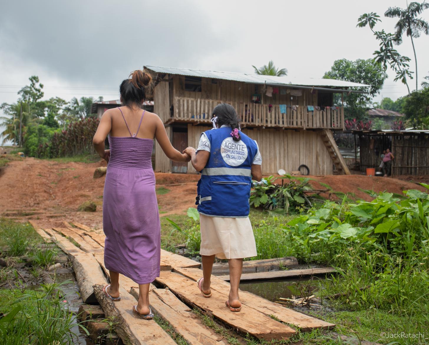 Dos mujeres caminando sobre unas tablas de madera en un espacio abierto lleno de vegetación tropical, y se dirigen hacia una edificación de dos plantas.