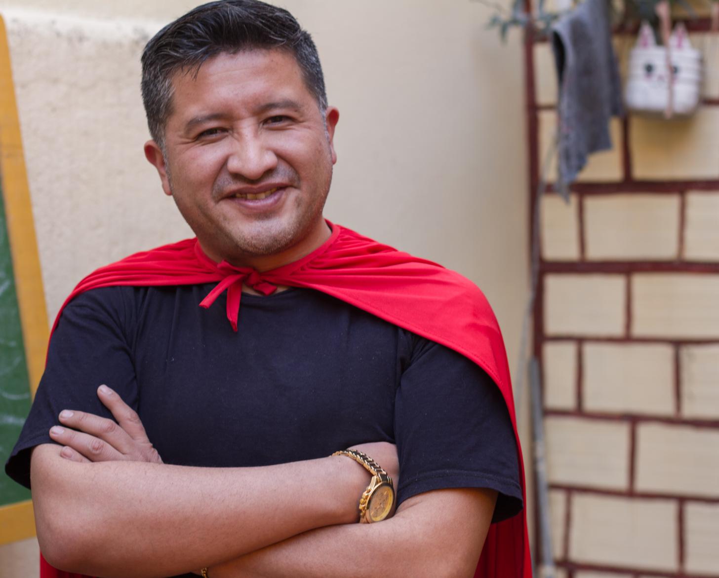 Un hombre sonriente frente a una pizarra, con anotaciones hechas con tiza blanca, y lleva puesta una capa roja de superhéroe.