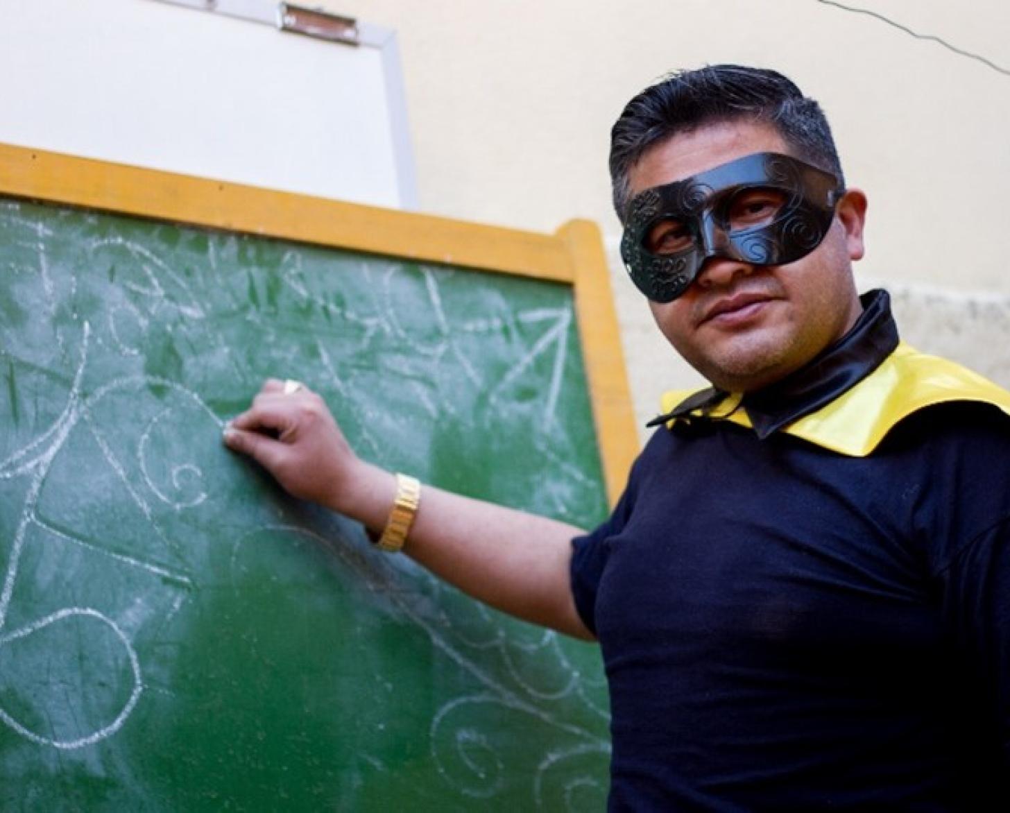 El súper profe boliviano disfrazado con una tiza en la mano frente a una pizarra.