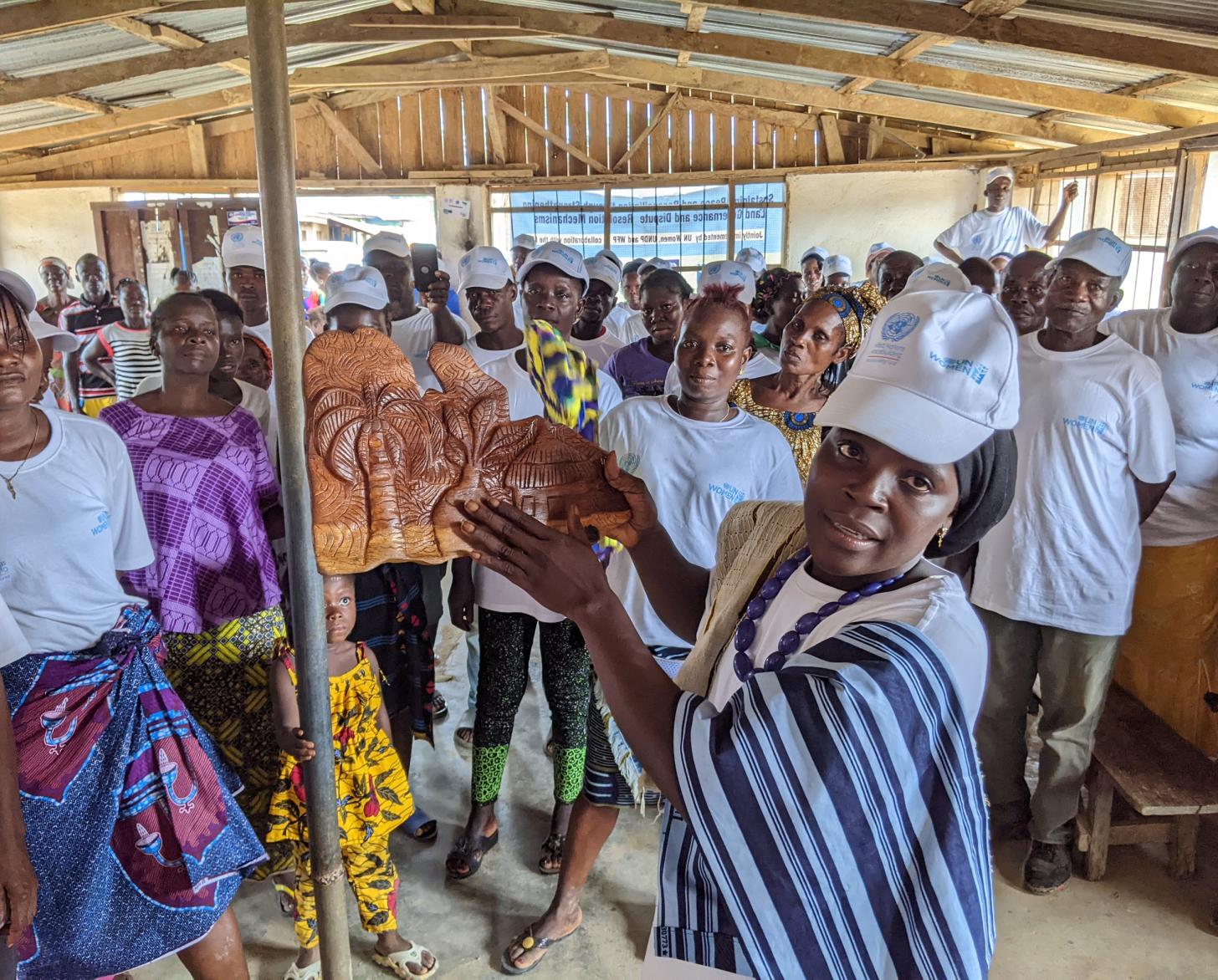 تجمع مجموعة من النساء والرجال في غرفة وينظرون نحو الكاميرا بينما تعرض إحدى النساء، في المقدمة، منحوتة مصنوعة يدويًا.