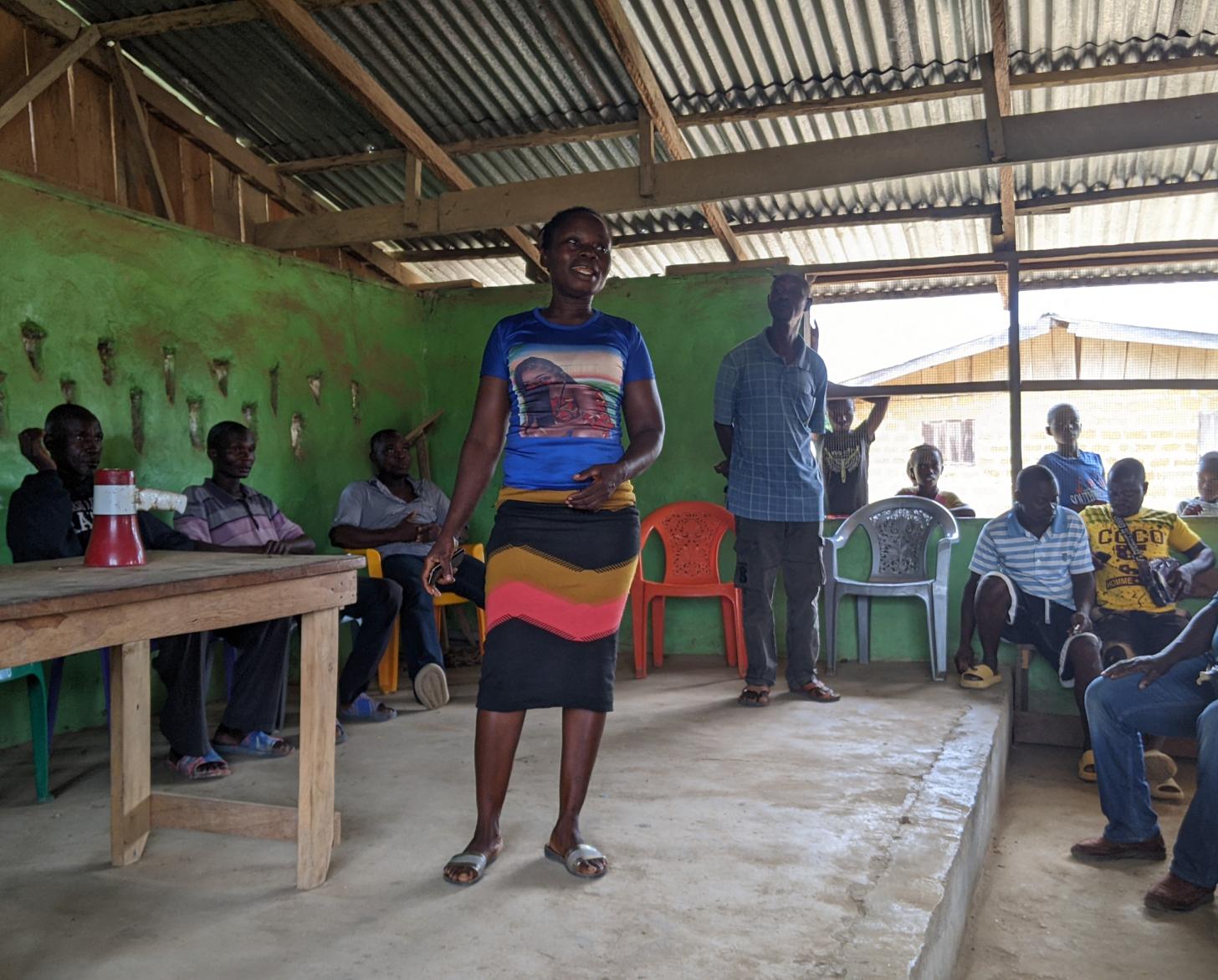 تقف امرأة على خشبة المسرح، محاطة بعدة أشخاص جالسين على الكراسي في غرفة ذات سقف مصنوع من الخشب والألواح المعدنية.