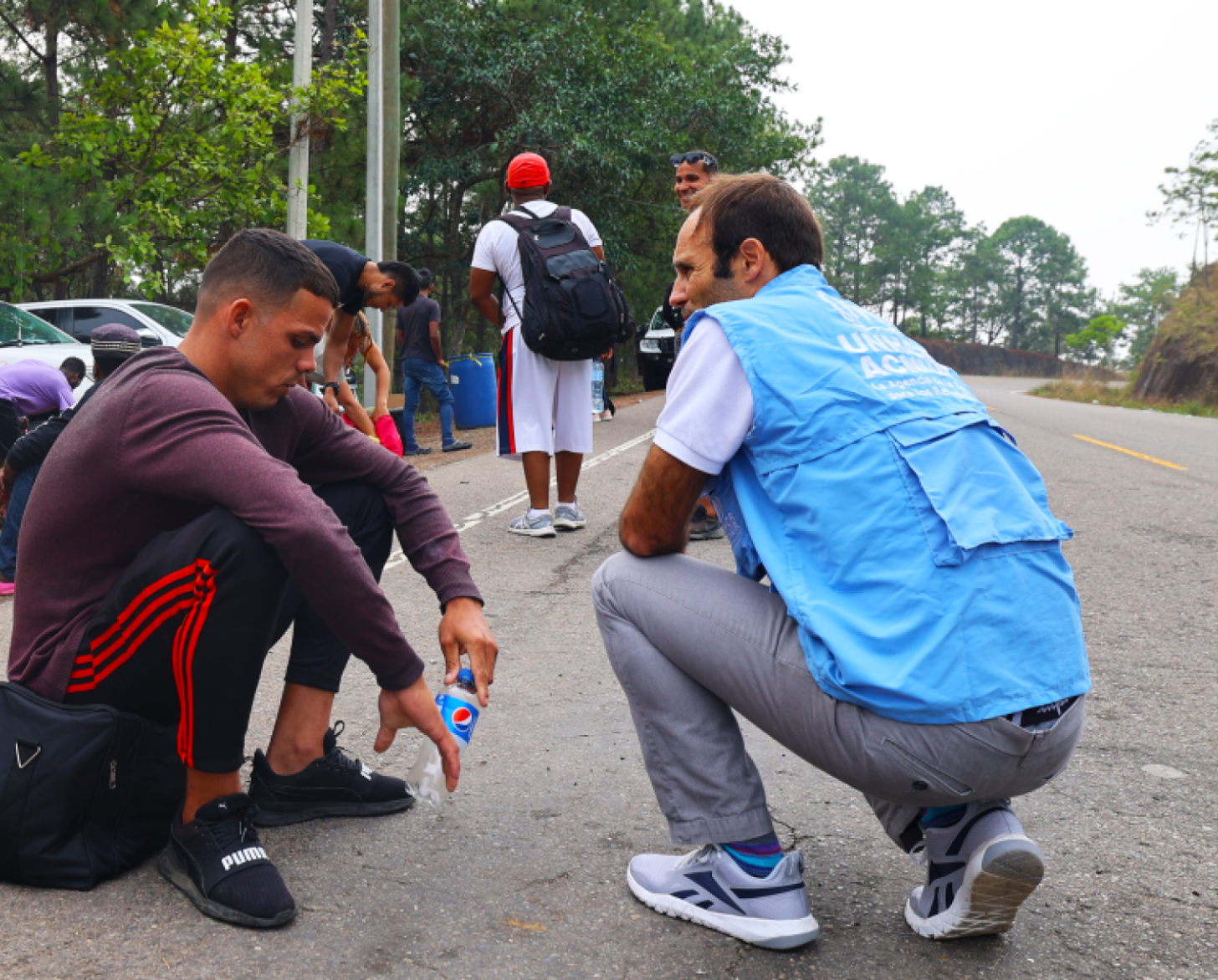 A man in a blue vest kneels on the ground next to another man crouched over, in a red shirt