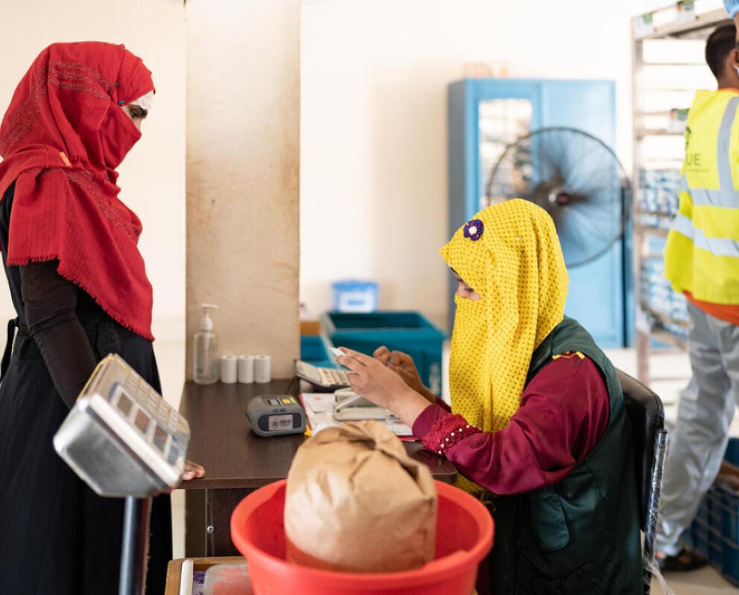 Woman in a a red veil and a black dress stands at a food distribution counter while a woman in a yellow veil and black dress weighs a bag on a weighing scale
