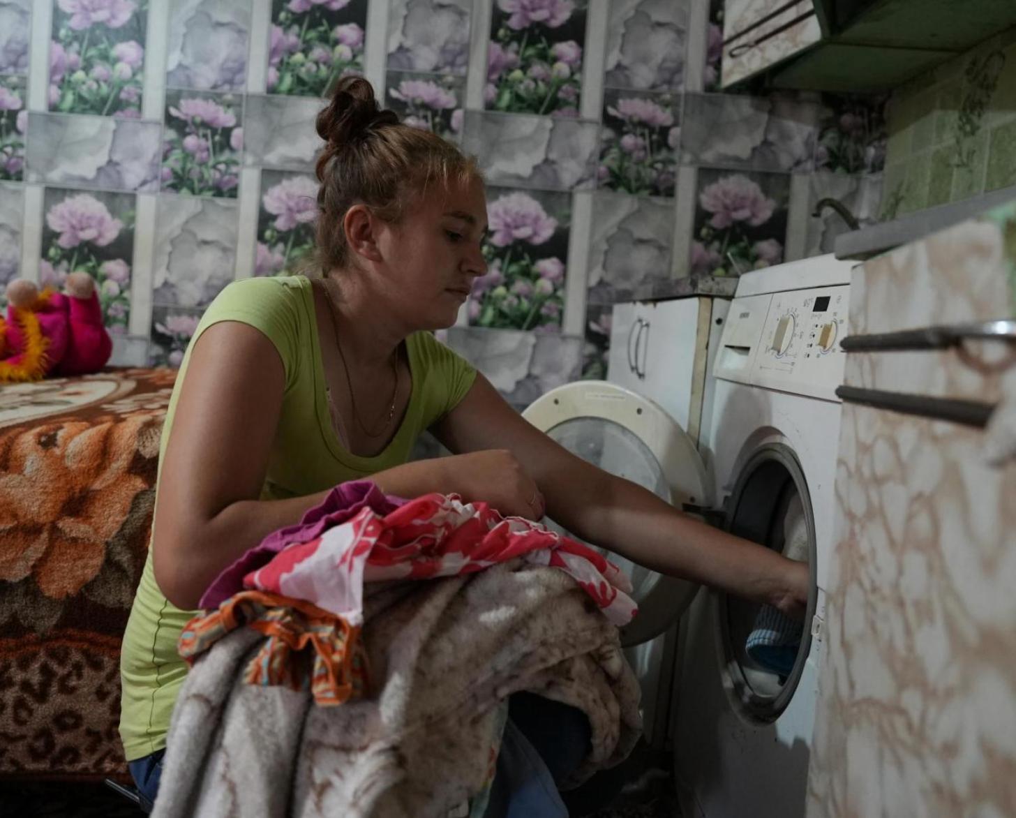 A woman puts clothes into a washing machine in a lightly-lit room.