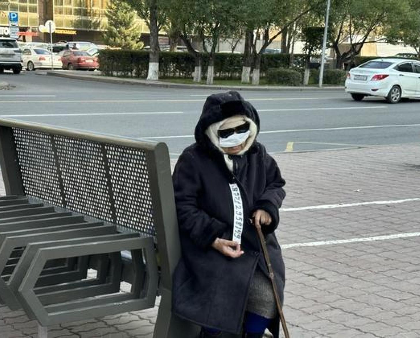 Женщина в повязке на лице и темных солнечных очках сидит на лавочке с тростью в руках