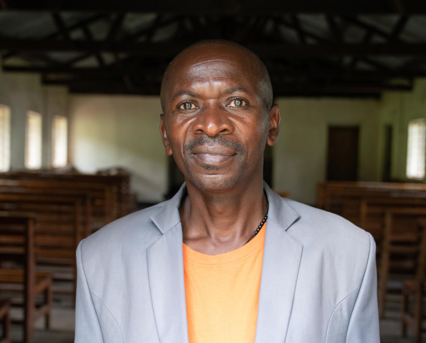 man in grey suit and orange top stands close to the camera, with a church interior behind him