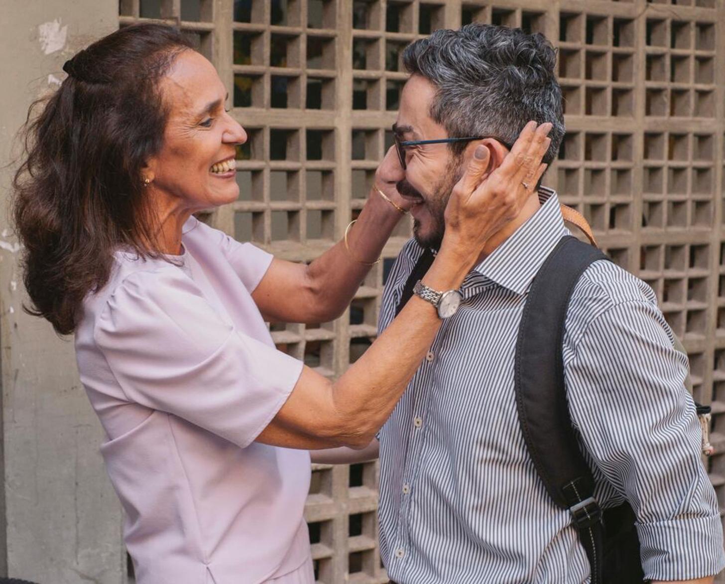 A woman in a pink shirt smiles and holds the face of a man in a grey shirt and glasses.