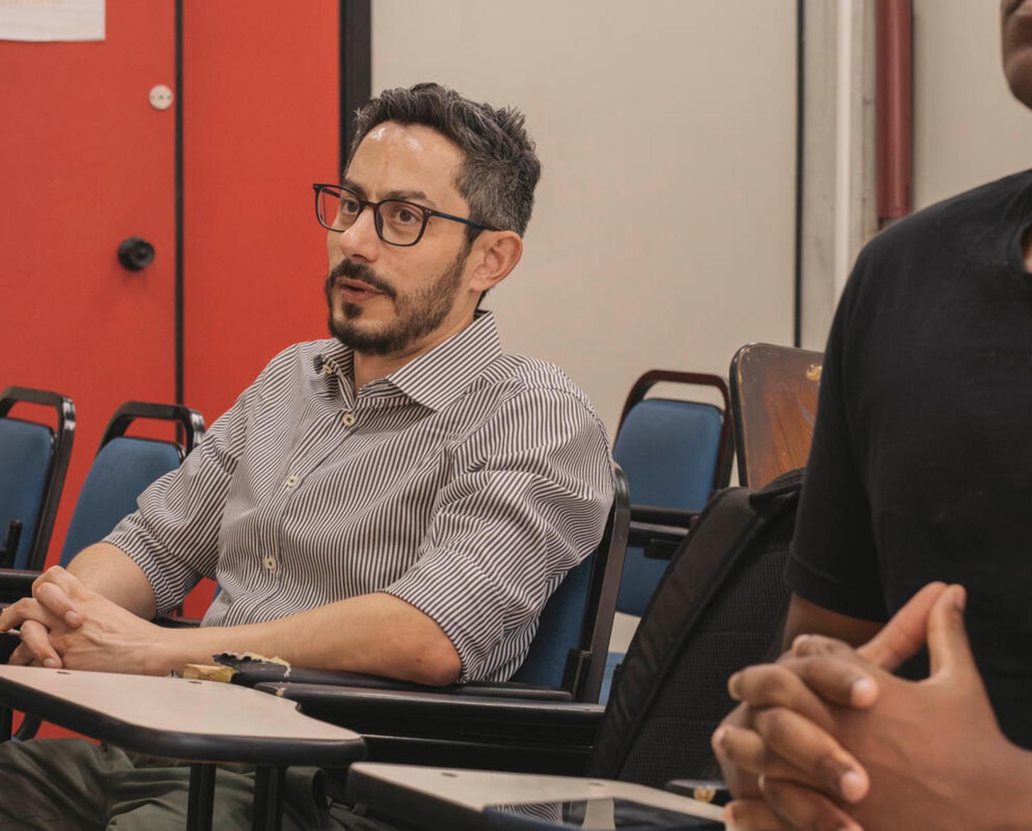 A man in a grey shirt and glasses sits at a desk in a classroom with bright orange walls behind him.