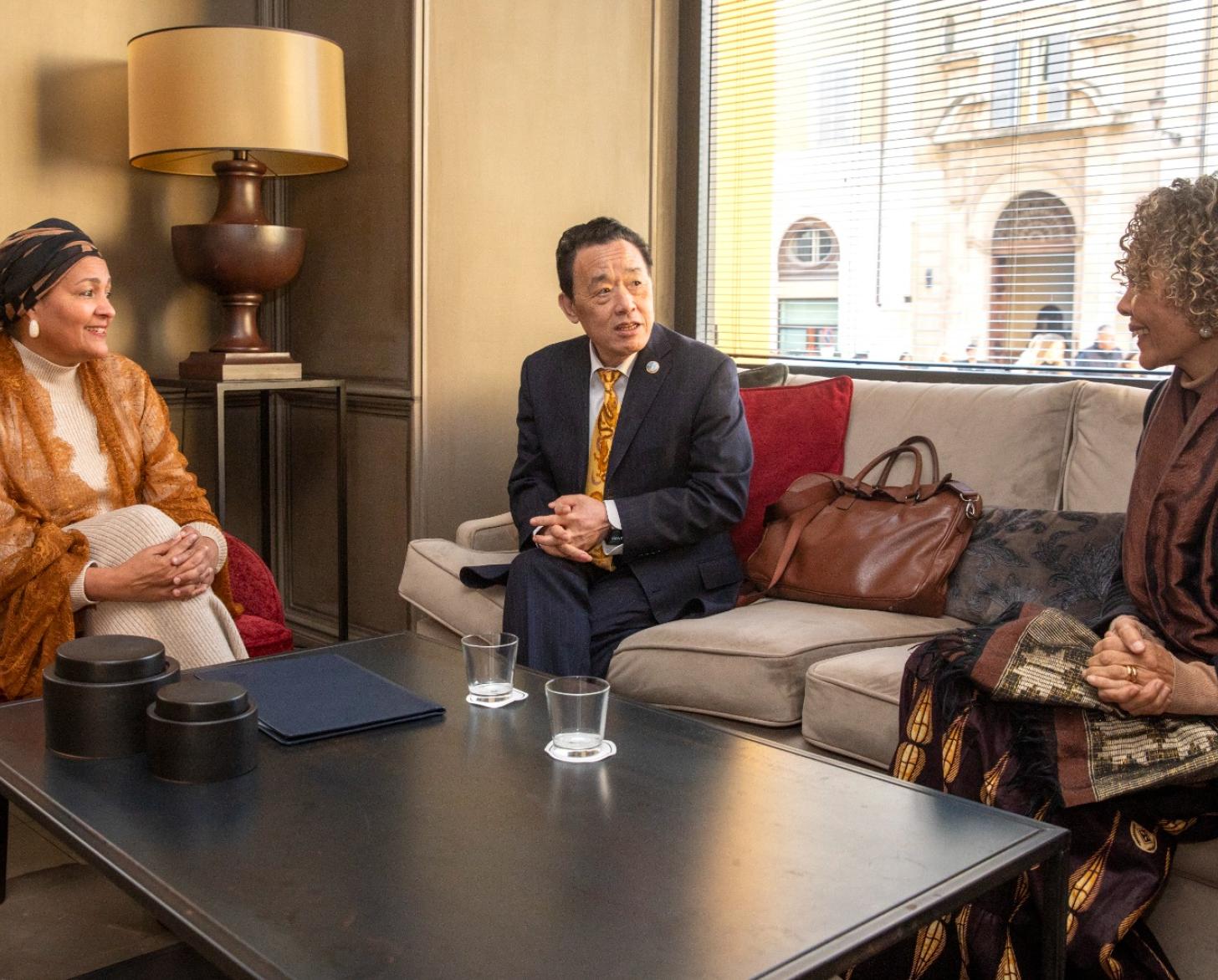 A woman in a lacey, brick-brown shawl and stylish headwrap (left) holds a meeting with two UN officials, all seated on couches in an informal setting.