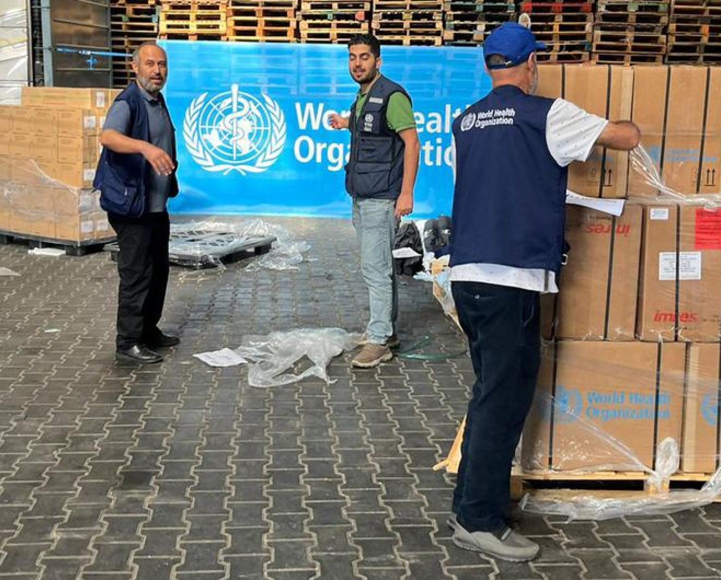Men unload boxes of supplies and boxes, and stand in front of a World Health Organization sign.