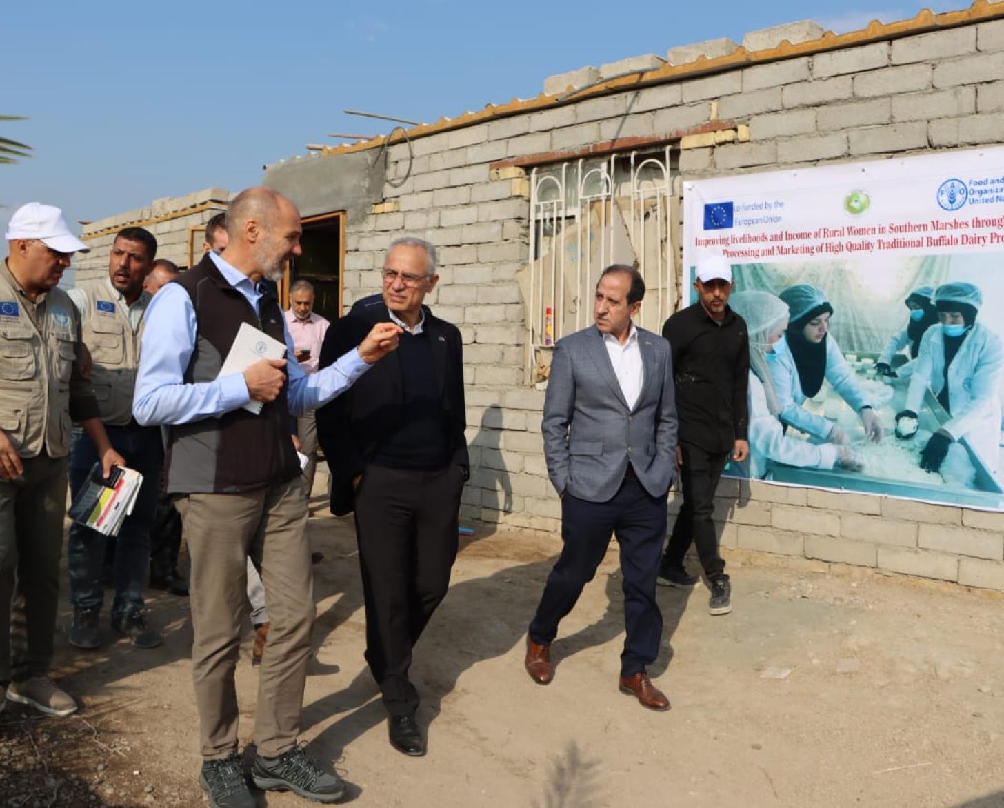 A group of men are walking on a dirt street. A building with exposed gray bricks is seen behind them.