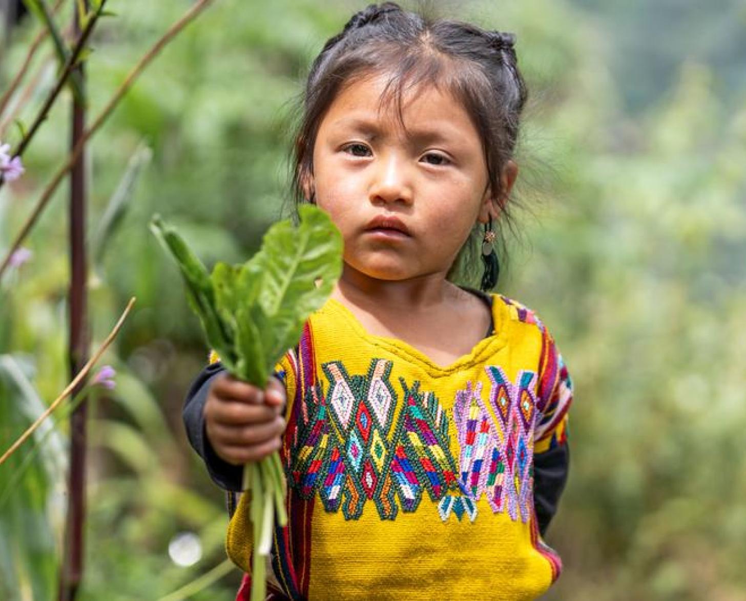 A young girl in a colorful shirt holds a handful of herbs towards the camera