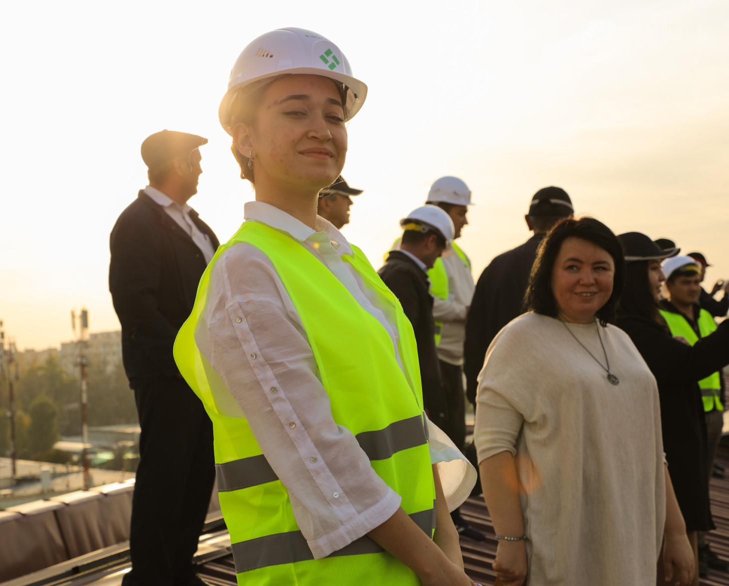 A woman in a green vest and hard hat smiles to the camera as other look on