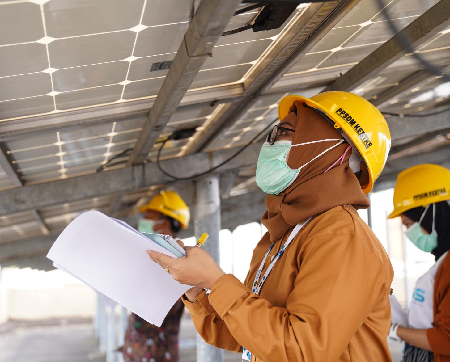 A woman in orange clothes and a hard hat stands under a solar panel