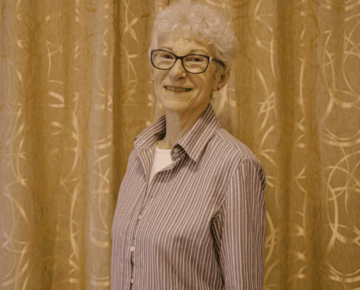 A pensioner in glasses and a striped shirt stands and smiles to the camera.