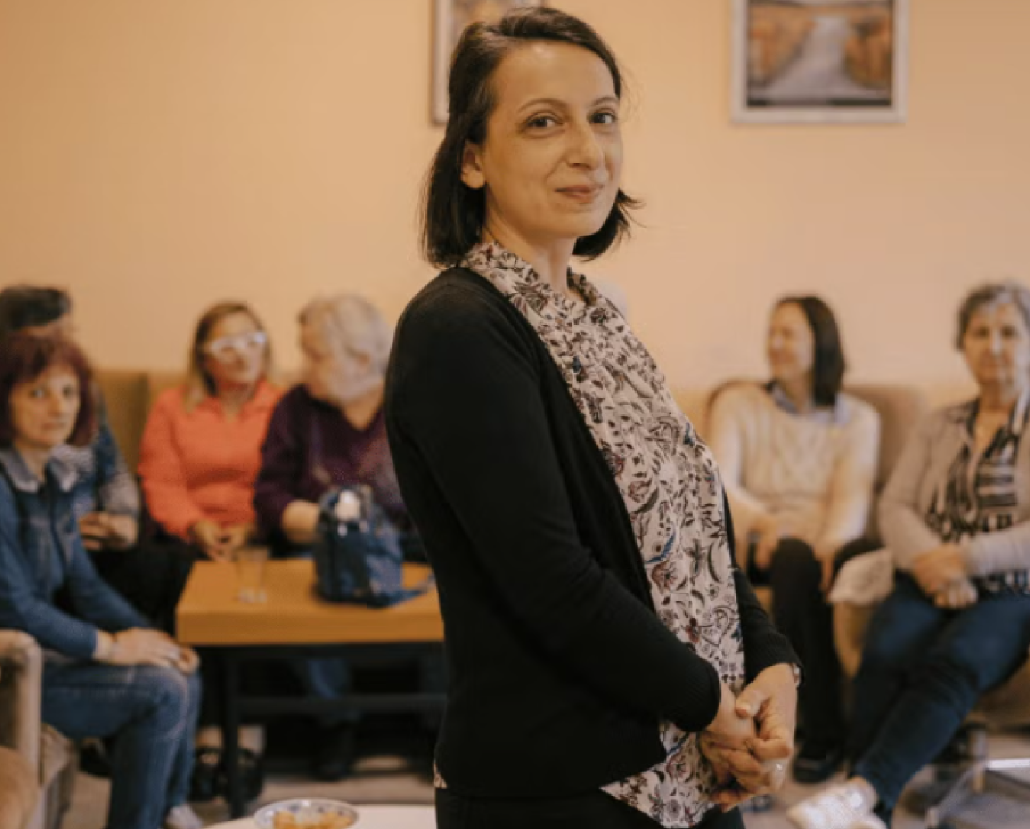 A pensioner stands and smiles to the camera, in the background a group of other pensioners are chatting amongst themselves.
