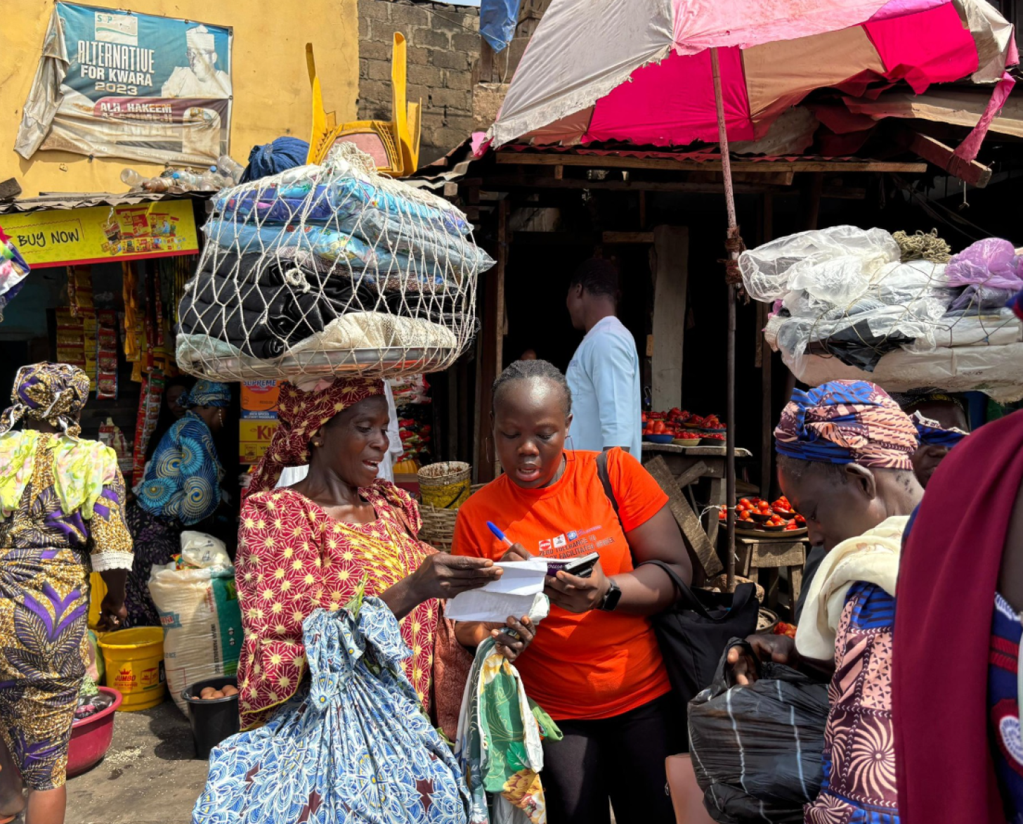 A Nigerian female volunteer stands in a busy marketplace and writes something down while talking to another woman.