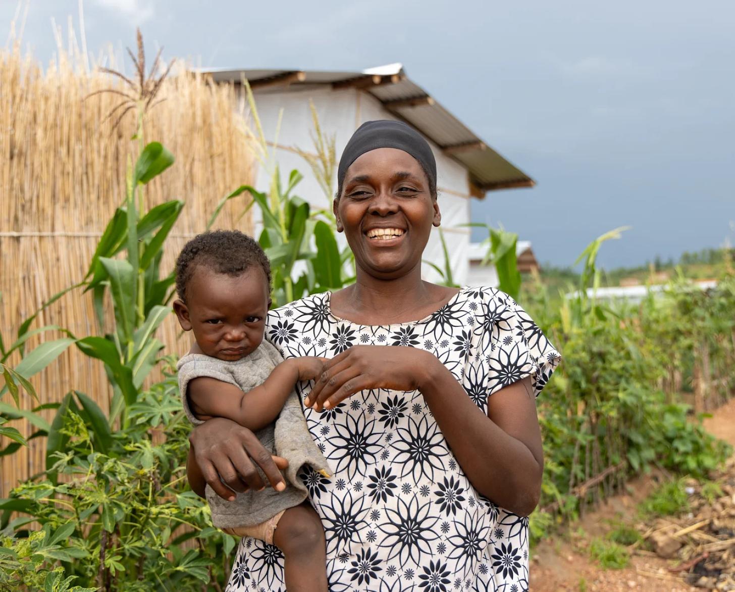 A mother in Burundi holds her baby and smiles in front of maize crops.