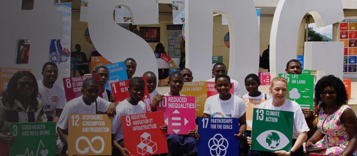 The Resident Coordinator of Cameroon smiles as she and young people kneel holding SDG signs.