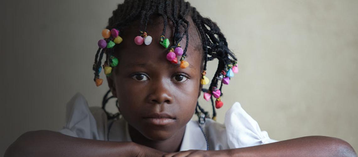 Una niña pequeña con su cabellera trenzada con coloridos accesorios, está sentada en su escritorio con las manos delante mientras mira directamente a la cámara.