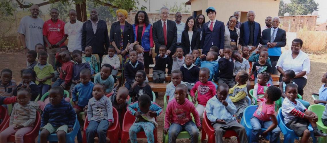 Children from a Neighbourhood Care Point (NCP) sit together with UN staff and Japan officials standing the background. 