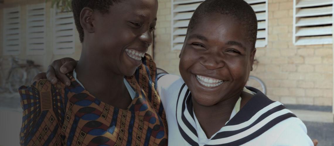 Two girls outside a building cheerfully smile with arms around each other. 