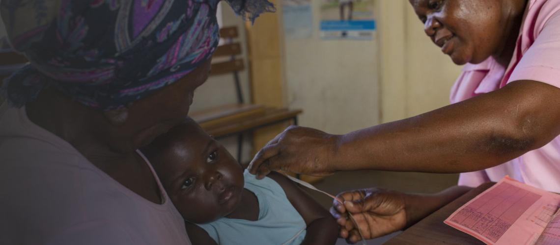 A woman holds a baby as a healthcare worker measures the child. 