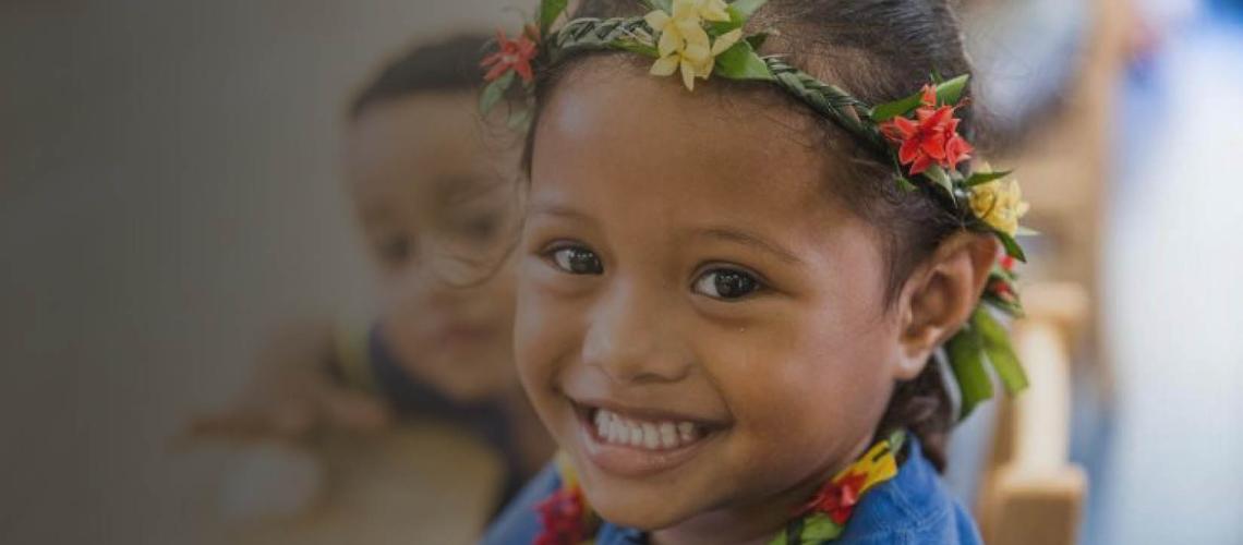 A little girl smiles happily at the camera as a little boy peeks behind her. 