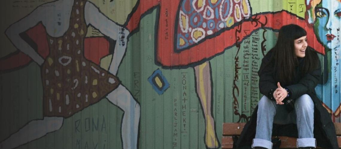 شابة تجلس على مقعد بالقرب من حائط مزيّن بالصور وتبتسم بسعادة لشخص خارج إطار الكاميرا.