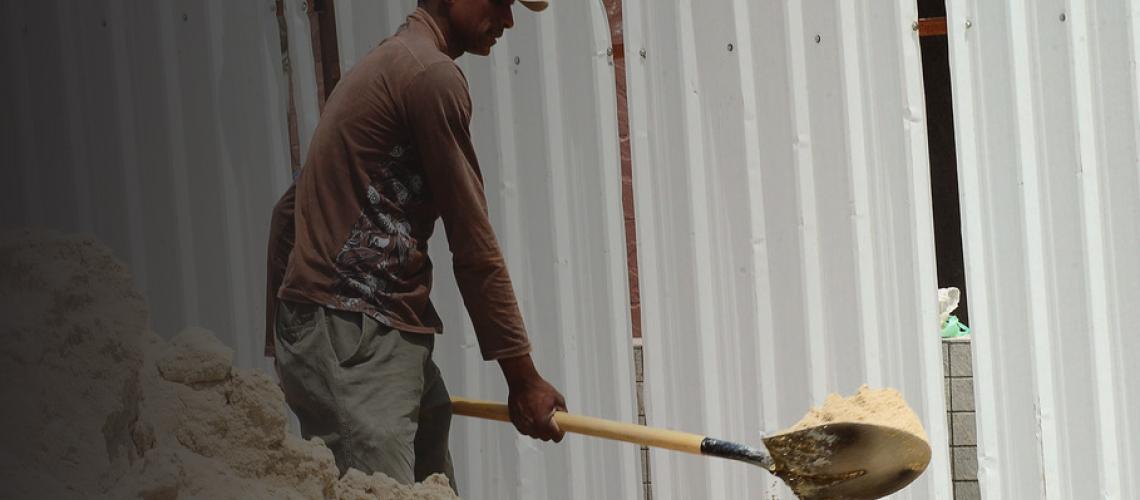 A man shovels sands from a large pile into a wheel barrow. 