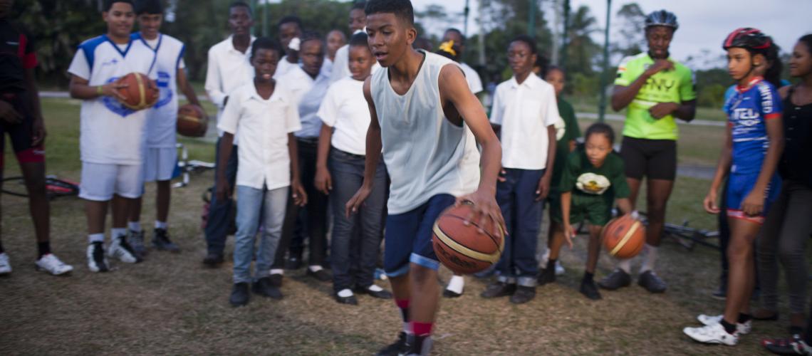 An adolescent boy is shown in the centre dribbling a basketball with other boys slightly blurred in the background cheering him on.
