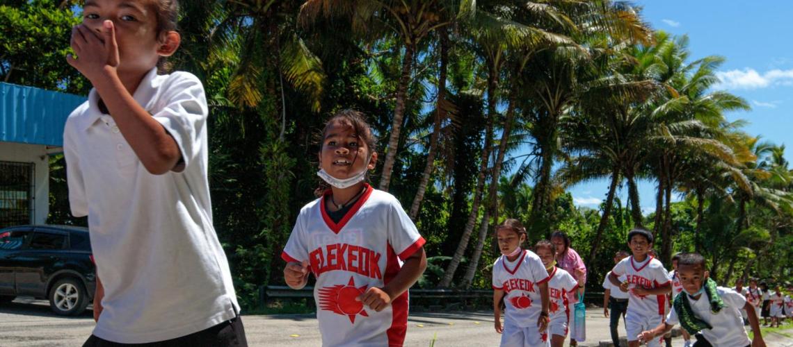 School children wearing uniforms walk in a single file line on a bright sunny day.
